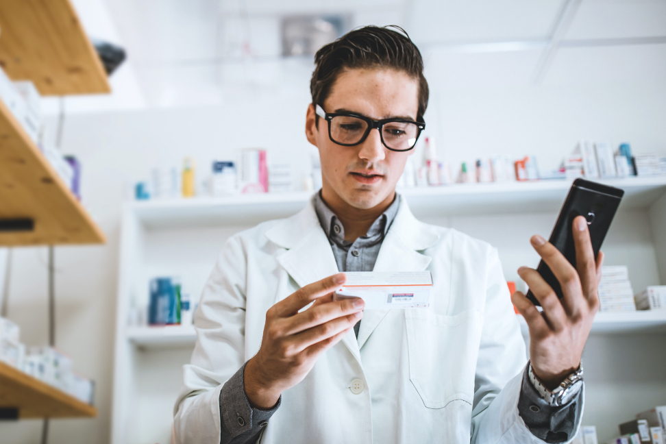 Männlicher Apotheker einer zugelassenen Apotheke von Unify Health, der mit seinem Mobiltelefon vor Regalen mit Medikamenten steht, um Paketdetails zu überprüfen