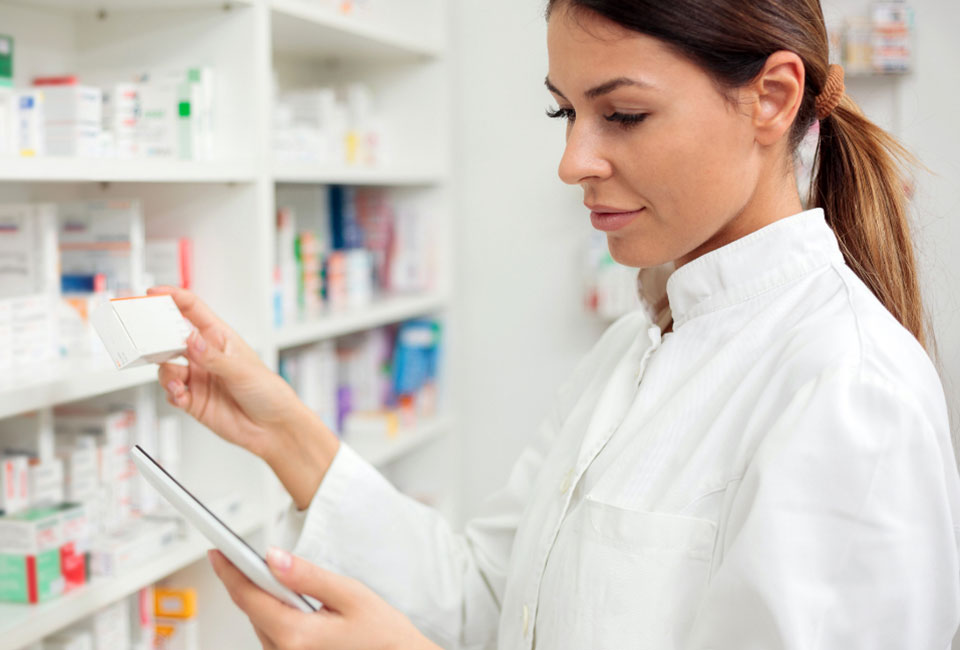 Young Mediterranean female pharmacist standing in front of shelves of medication checking packet detail while holding iPad