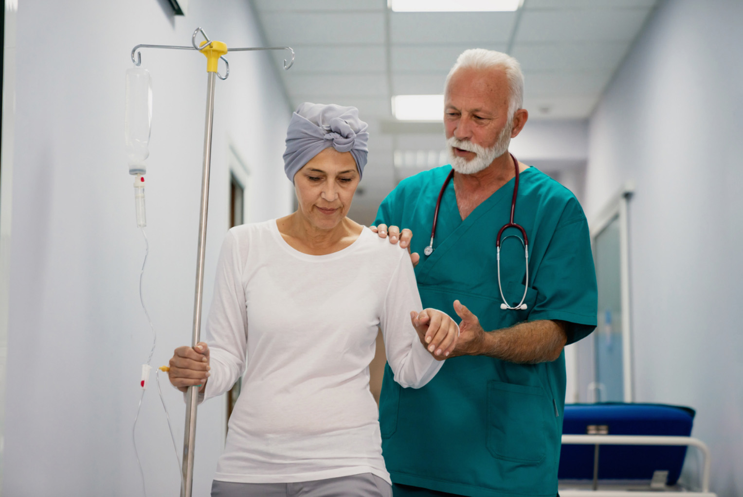 Older male nurse walking through a hospital corridor helping and holding hand of middle-aged hispanic female patient on infusion