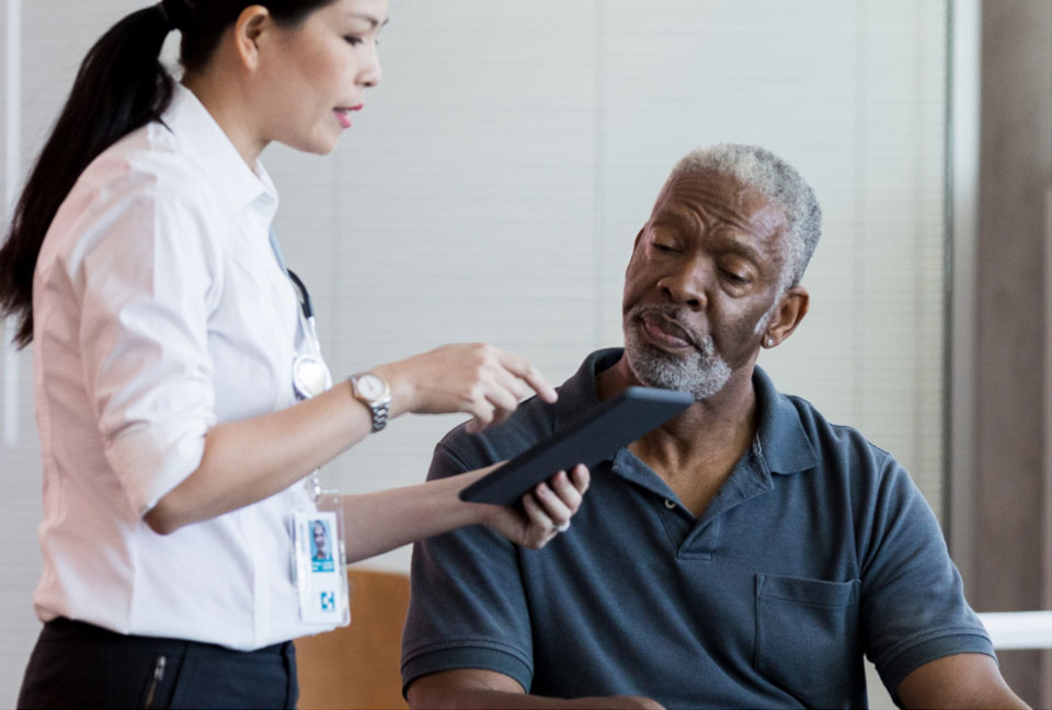 Middle aged white male doctor sitting at his desk holding a iPad and pointing out information to a middle aged white female patient