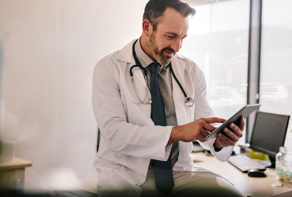 Male Unify Health doctor sitting on his desk with his computer in the background holding and pointing at his iPad